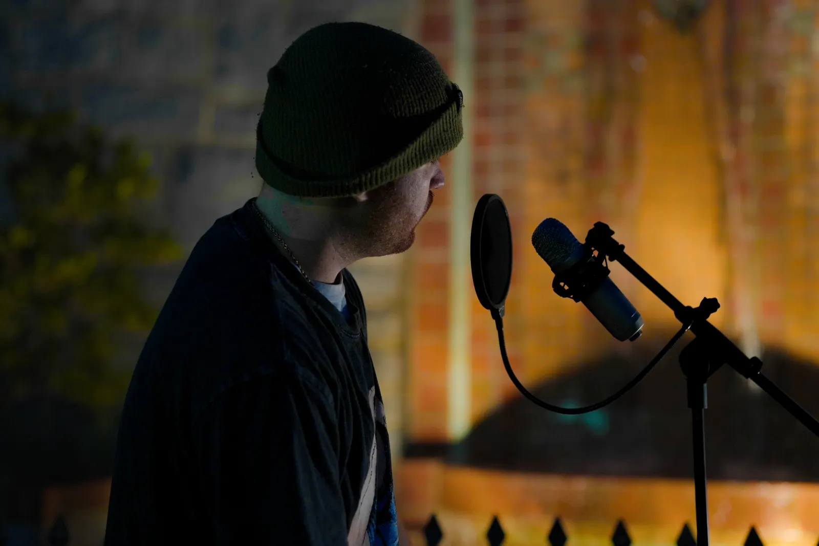 A vocalist in a beanie, in profile, singing into a condenser microphone at night